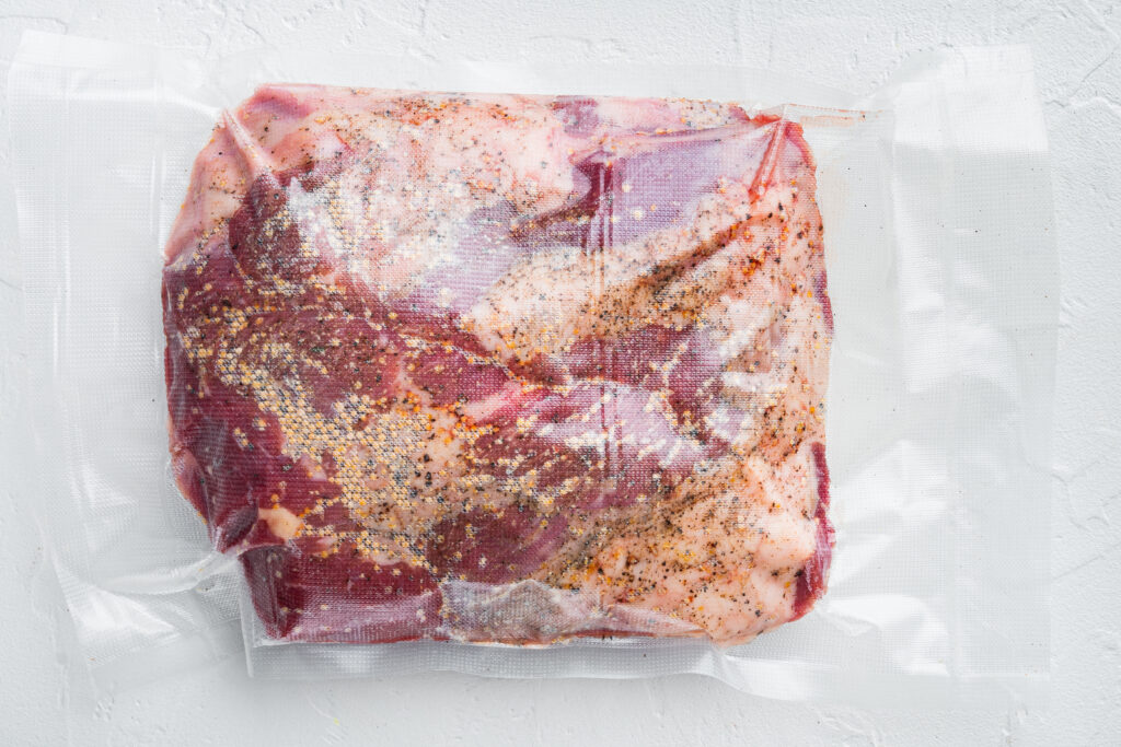 Raw steak with pepper and herbs in a vacuum-sealed bag on a white surface.