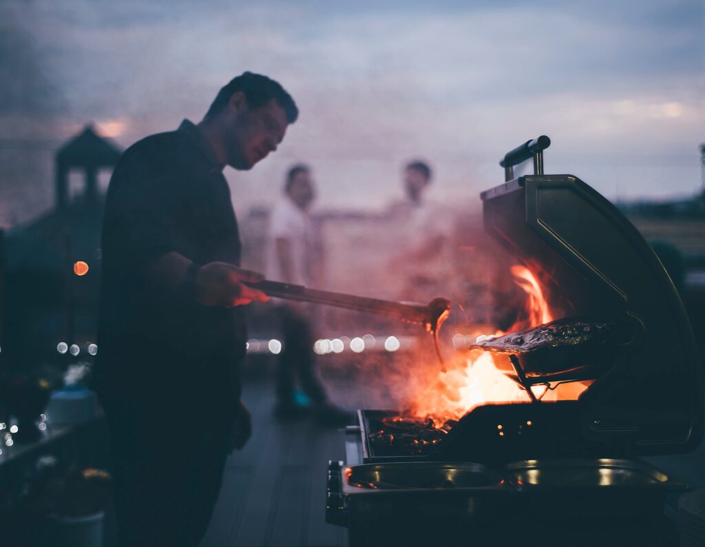 Man tending a glowing charcoal grill on a deck at dusk, with others blurred in the background.