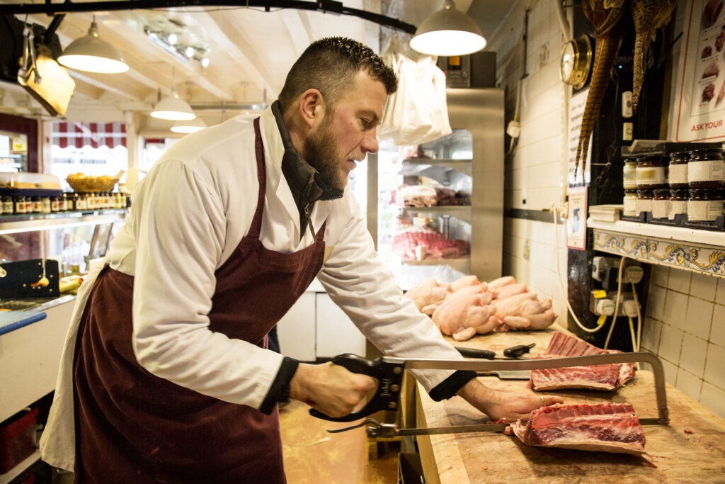Butcher in a white coat and burgundy apron slices a ribeye with a handsaw in a meat counter.