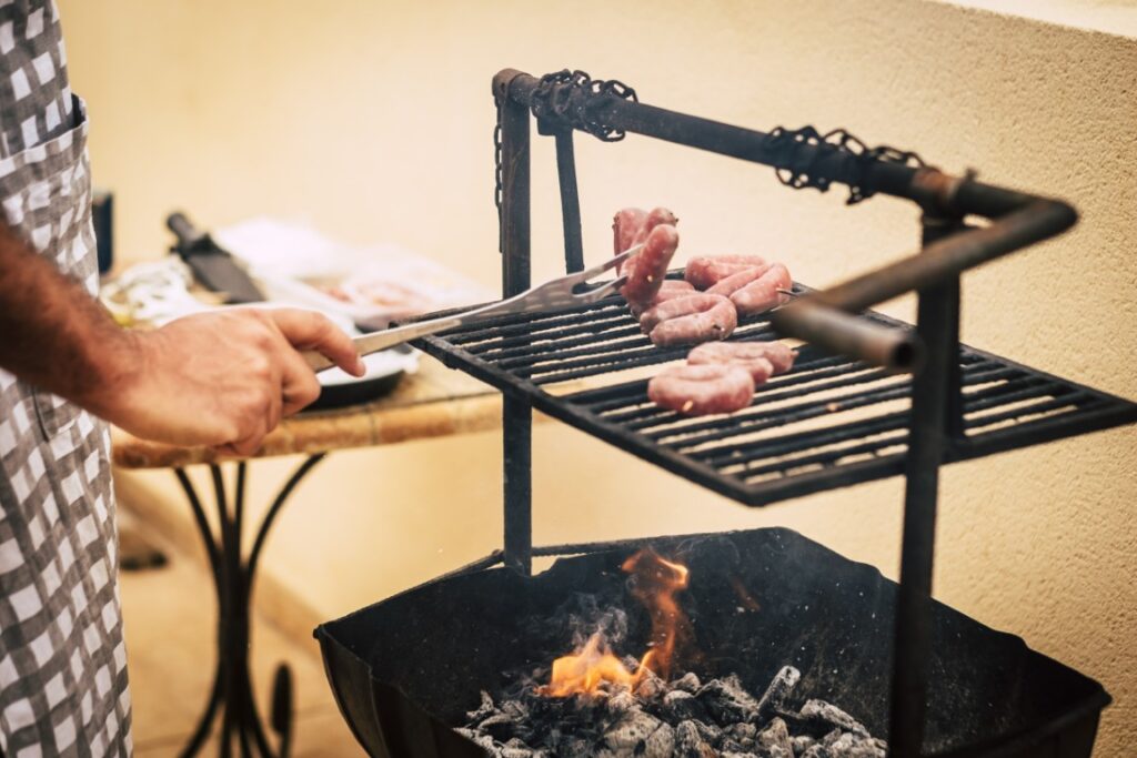 Person in a checkered shirt uses tongs to turn raw sausages on a charcoal grill over glowing coals in a small outdoor setup.