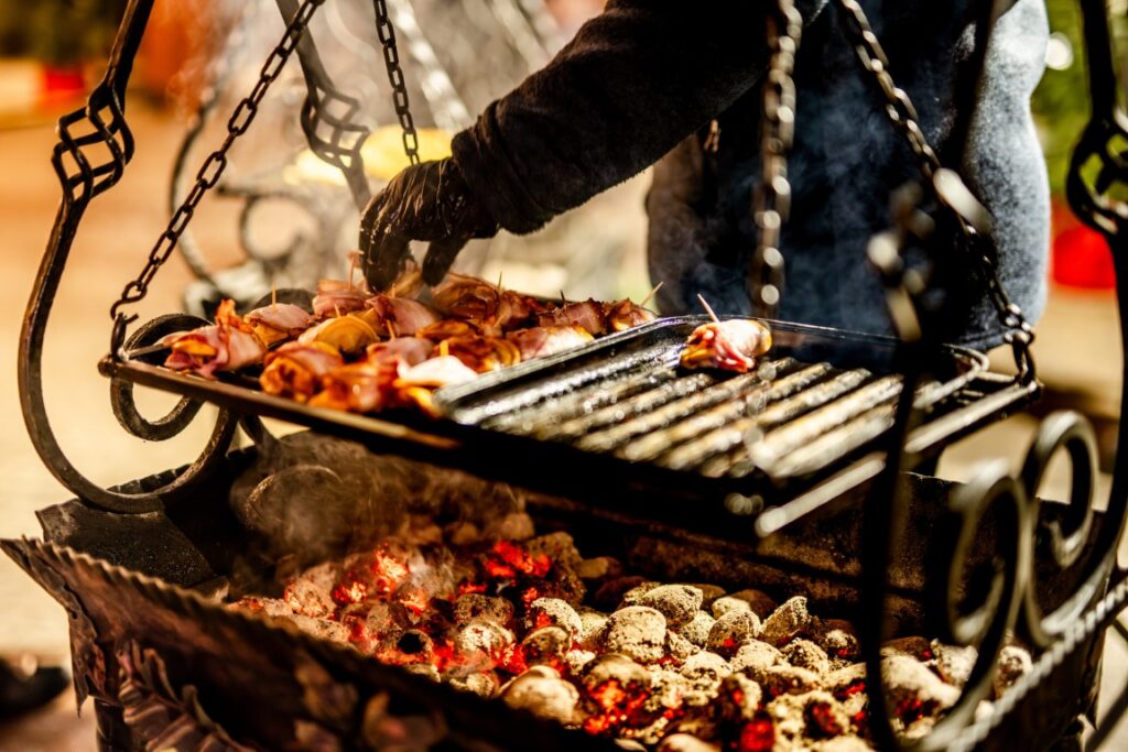 Person wearing black gloves grilling skewered meat over glowing charcoal in an ornate outdoor grill.