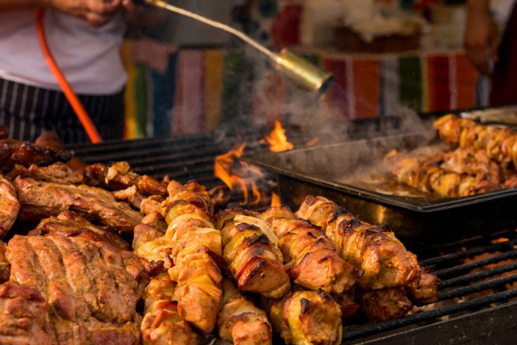 Skewers of grilled meat on a sizzling open barbecue with visible flames and a colorful stall backdrop behind.