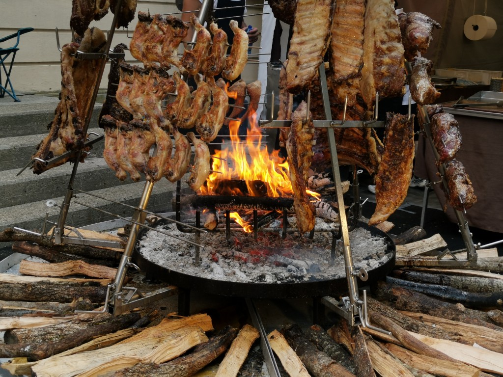 Rotisserie-style barbecue with multiple racks of meat cooking over an open flame, surrounded by logs and a bed of hot coals.
