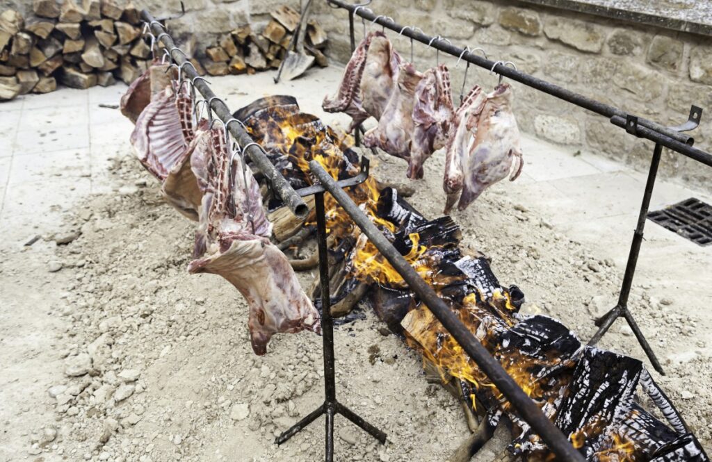 Raw meat cuts hung on metal racks above a wood fire outdoors, with a stone wall in the background.