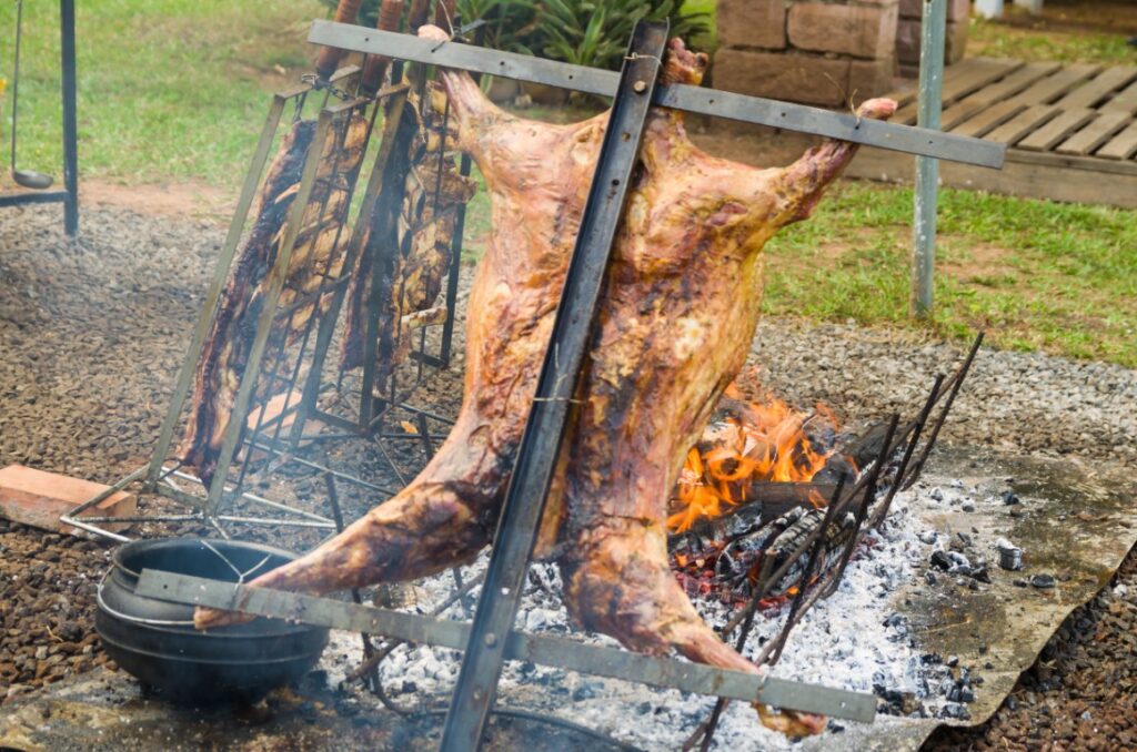 Two halves of a carcass being spit-roasted over a large outdoor fire, supported by metal frames in a backyard setting.