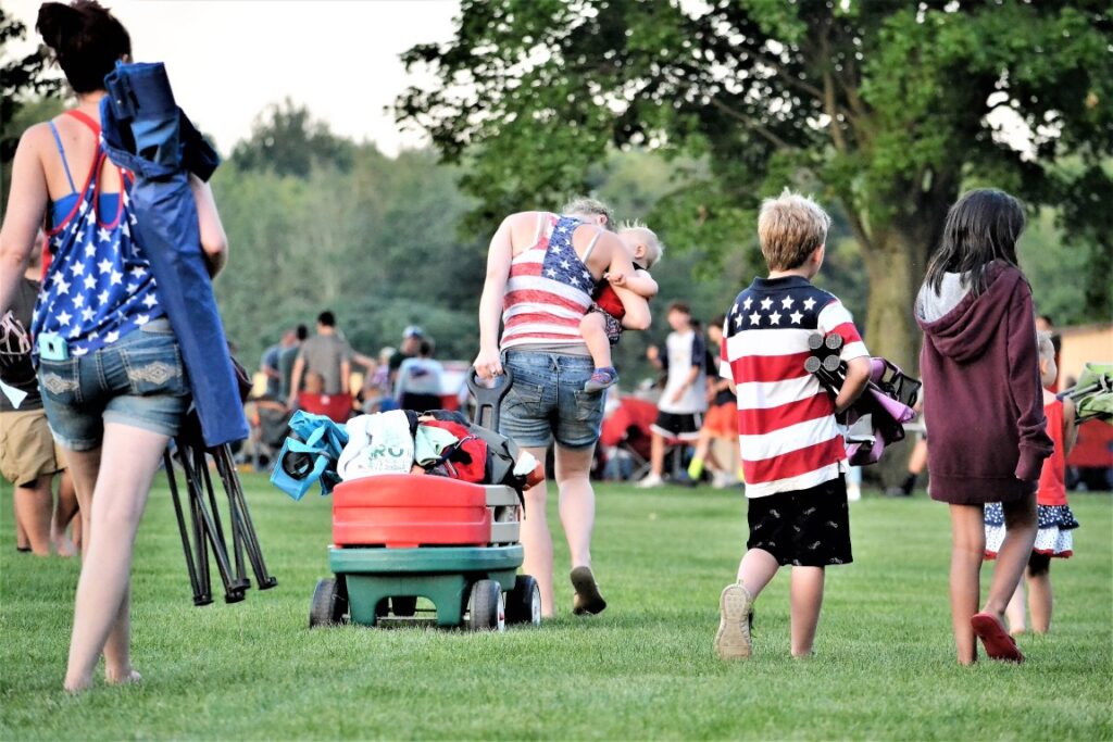 SBBlood Pressure 6 People at a park gathering on a sunny day; a woman carries a baby as others walk by with a wagon loaded with bags and gear, wearing patriotic outfits.