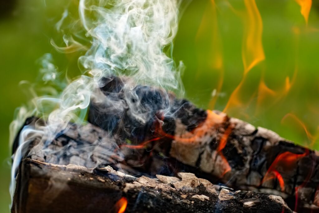 Log with glowing embers and curling white smoke rising from a campfire-like flame on charred wood