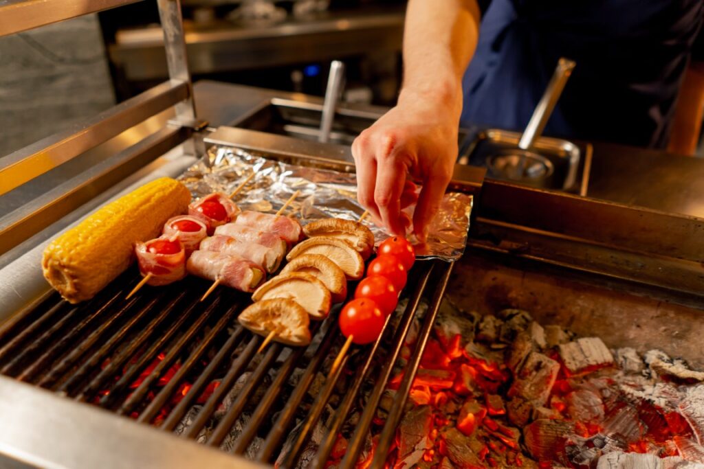 Hand placing bacon-wrapped skewers, mushrooms, and cherry tomatoes on a charcoal grill beside a corn cob drying nearby on foil-lined rack.
