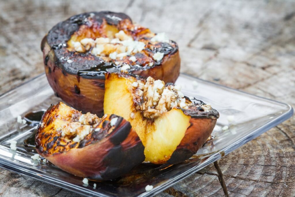 Grilled peach halves with chocolate glaze and crumb topping on a clear plate outdoors.