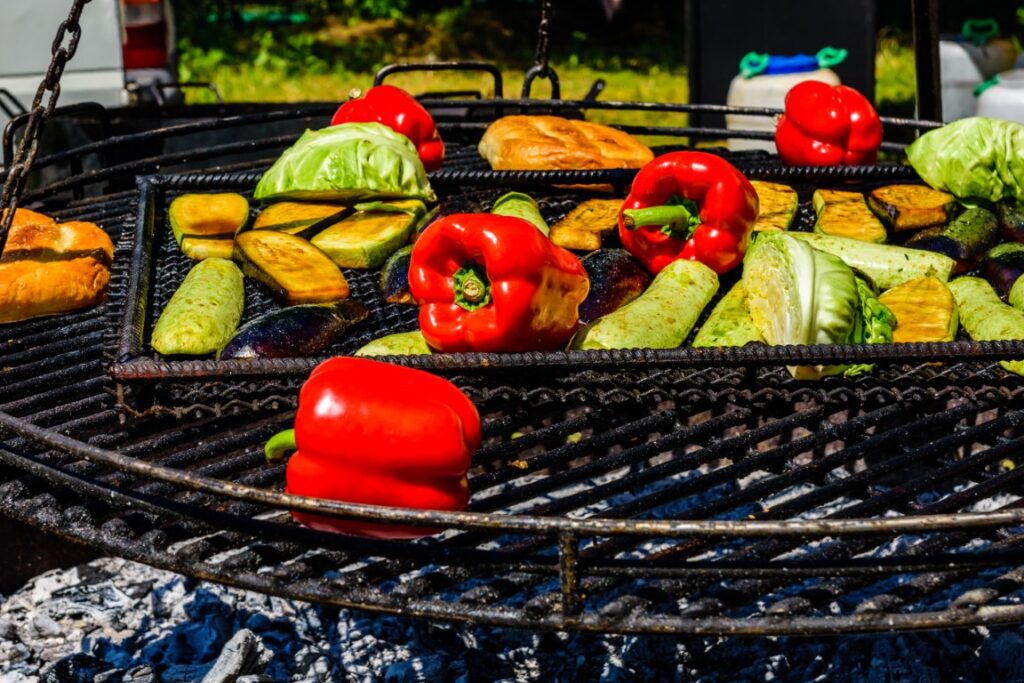 Assorted vegetables, including red bell peppers, zucchini, cabbage, and eggplant, grilling on a barbecue over hot coals.