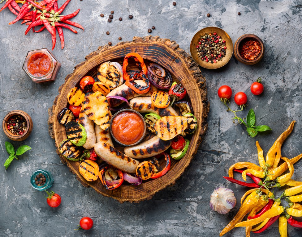 Assorted grilled sausages and vegetables on a rustic wooden platter with tomato dip, surrounded by chili peppers, garlic, and fresh herbs.