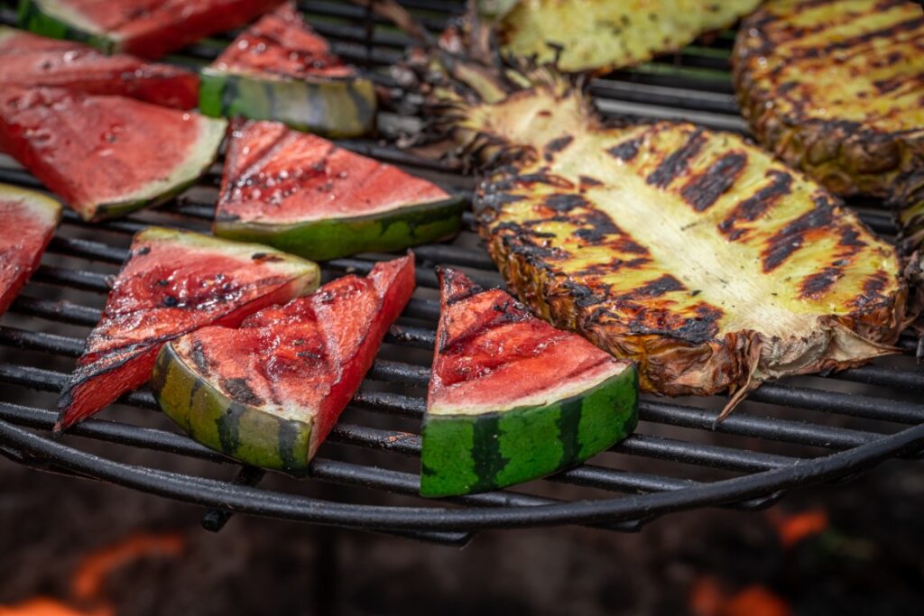 Watermelon wedges grilling on a barbecue with pineapple slices in the background.