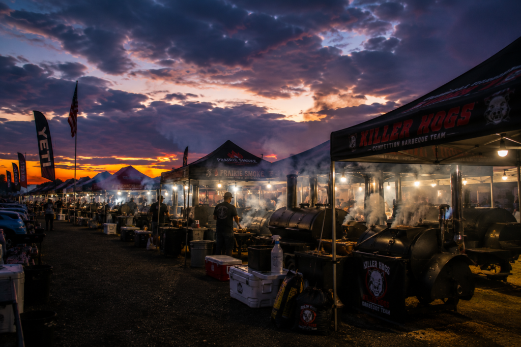 Row of barbecue stalls at dusk with smokers, tents, and a glowing sunset sky behind; smoke rises from grills.