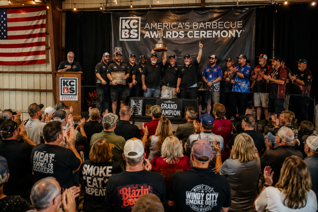 Group of men on stage at the KCBS America's Barbecue Awards Ceremony, one holding the tall trophy aloft while others cheer. Audience claps in a large hall with an American flag visible on the side.