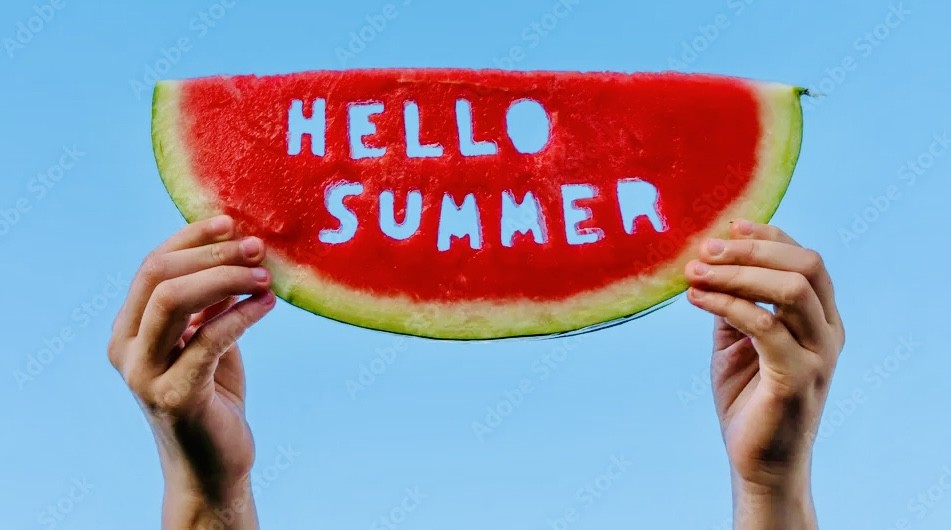 Two hands hold up a large slice of watermelon with the words 'HELLO SUMMER' written on the red flesh against a bright blue sky.
