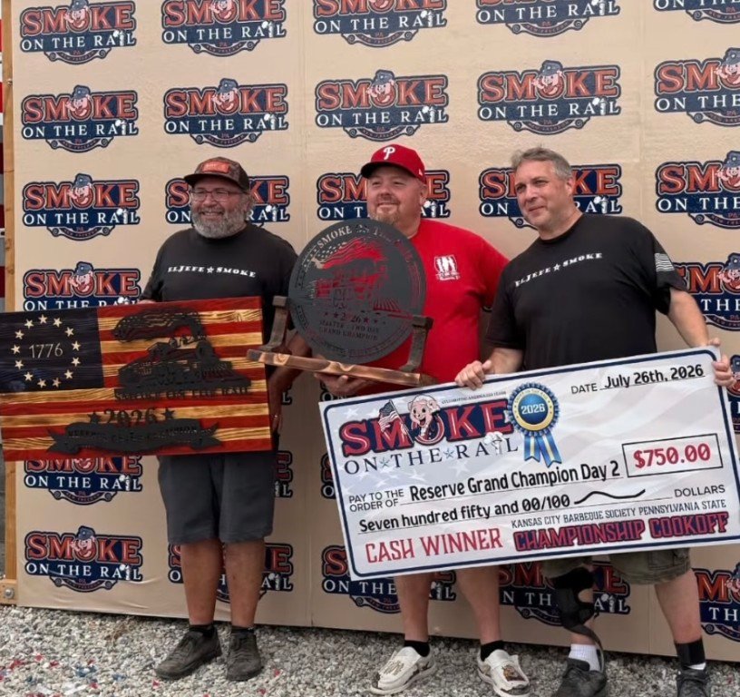 Screenshot Three men pose with awards at Smoke on the Rail; left holds a wooden American flag plaque, center holds a circular metal trophy, right holds a large $750 prize check (Reserve Grand Champion Day 2).