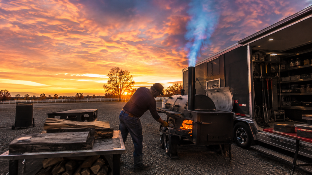 Smoke_Clears 1 - BBQ News Man tending a wood-fired smoker attached to a trailer at sunset on a gravel lot, with flames visible in the firebox.